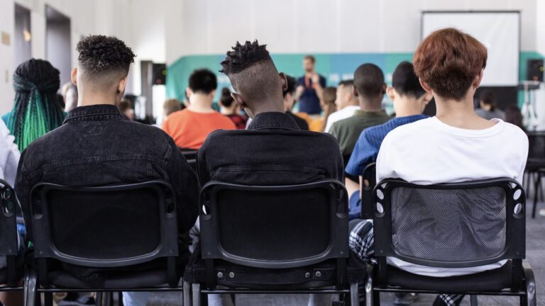three young men with their backs to the camera sitting in a class as the teacher takes a class