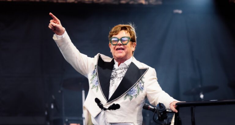 Elton John performing at a piano, his arm raised looking at the crowd.