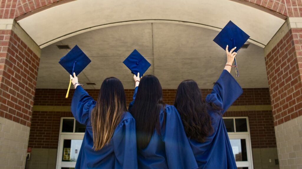three graduates in gowns holding up their dark blue graduation caps