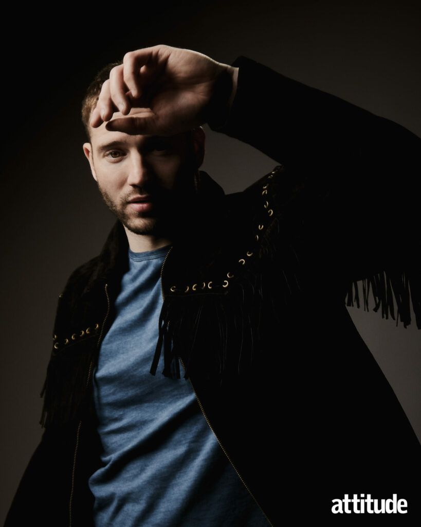 Alexander Lincoln in a blue t-shirt, a black cowboy-like jacket with his arm raised.