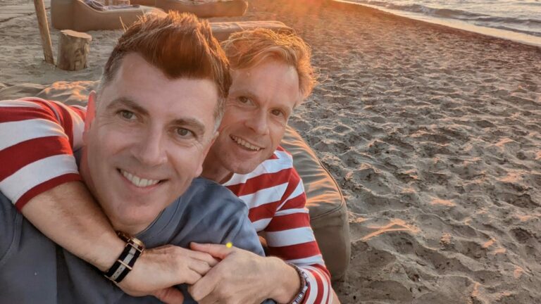 Two men hug on a beach with sunset in the background