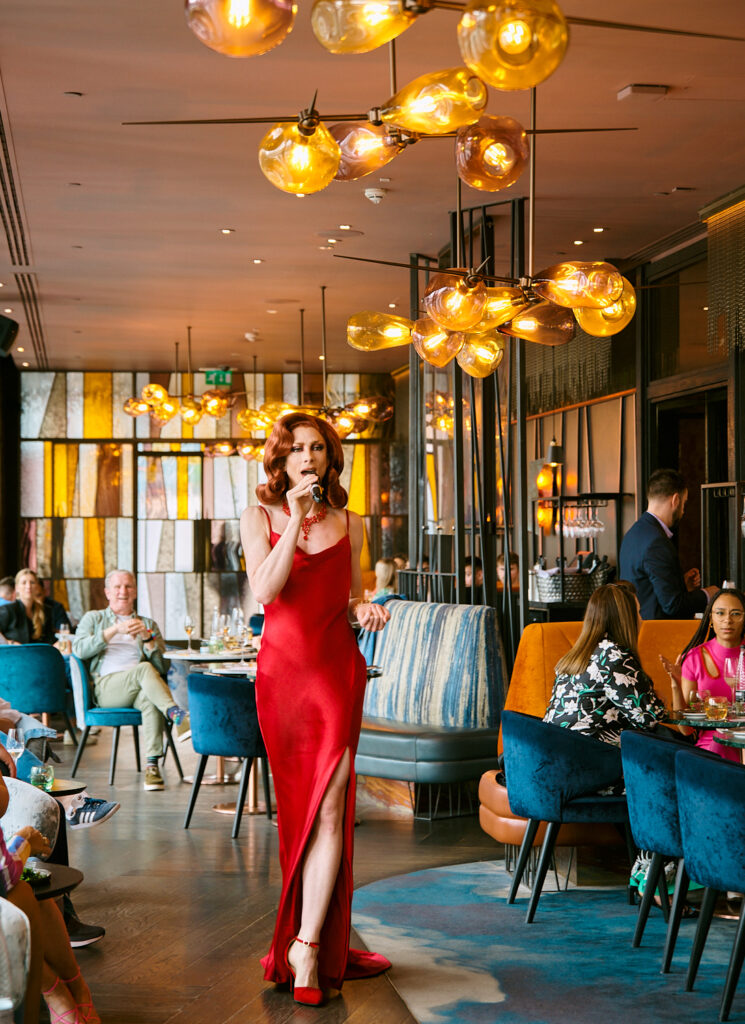 Drag Queen Sue Gives a F*ck is performing in a restaurant space. She wears a red slim ballgown, red high heels and red hair.