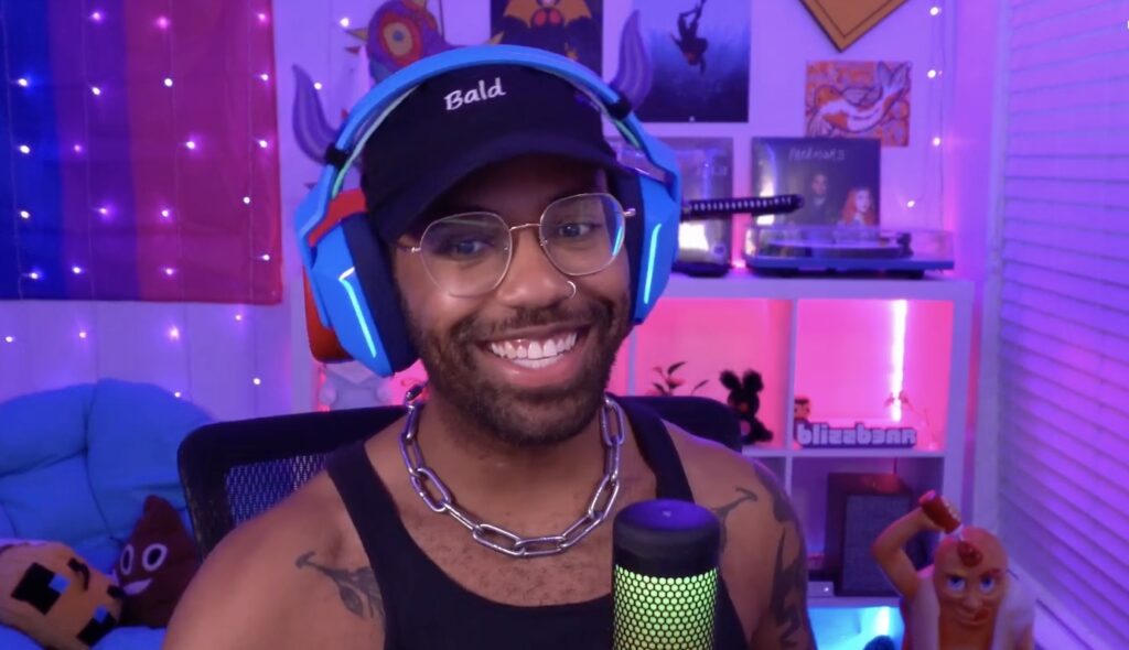 Blizzb3ar in his stream set up, wearing blue headphones, a cap, silver chain and black vest