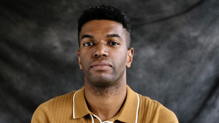 Okechukwu Nzelu sat against a misty grey background wearing a mustard top.