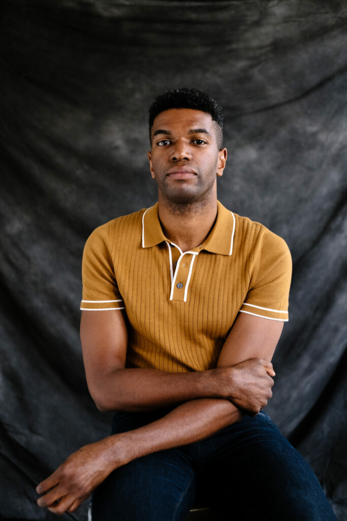 Okechukwu Nzelu sat against a misty grey background wearing a mustard top.