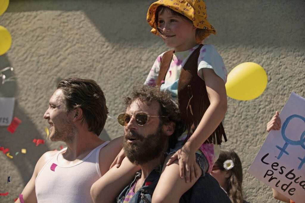 FAIRYLAND still. Two men, one in a white vest and the other a darker shirt. The latter has a young girl on his shoulders. She's wearing a yellow hat and tie-dye top.