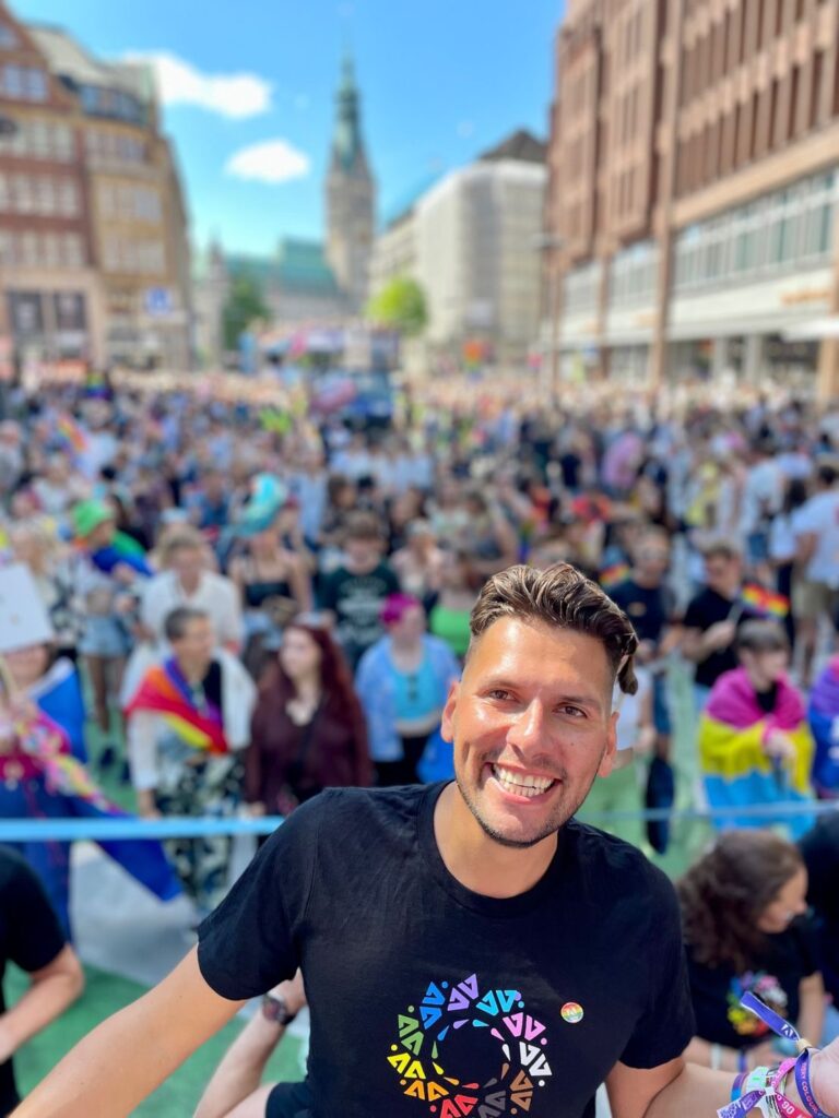 A man stands smiling at the camera in front of a crowd at a Pride event