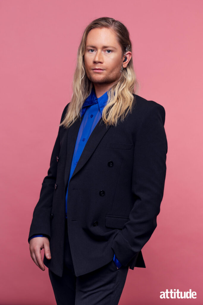 Luke Christian standing in a black suit, blue shirt against a pink background.