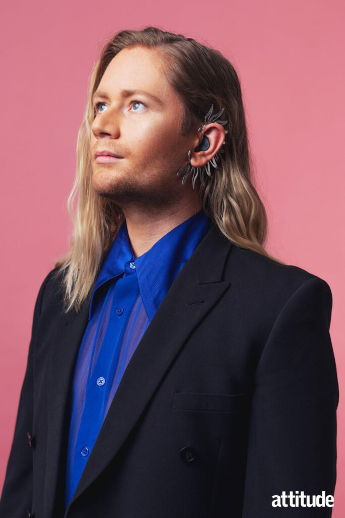 Luke Christian standing in a black suit, blue shirt against a pink background.