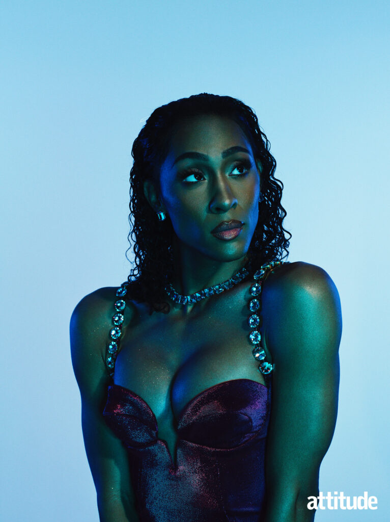 Michaela Jaé Rodriguez looking up to the right against a pale blue background. She wearing a deep purple dress with gems.