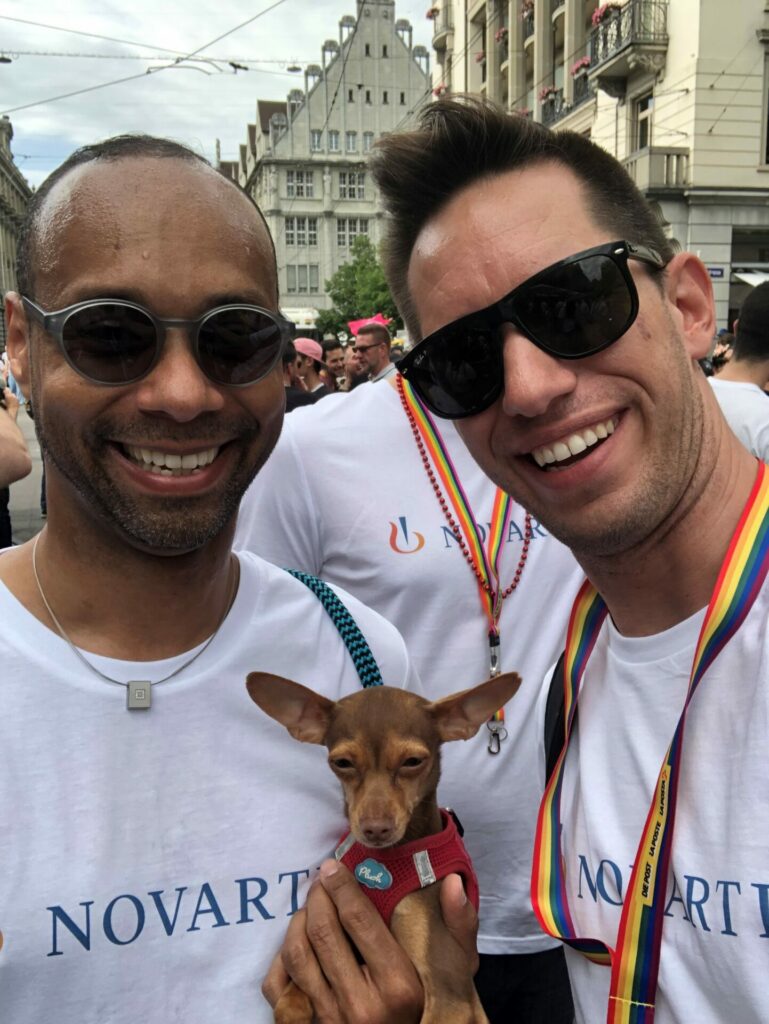 Two men wearing white T-shirts with rainbow lanyards around their neck hold a small brown dog