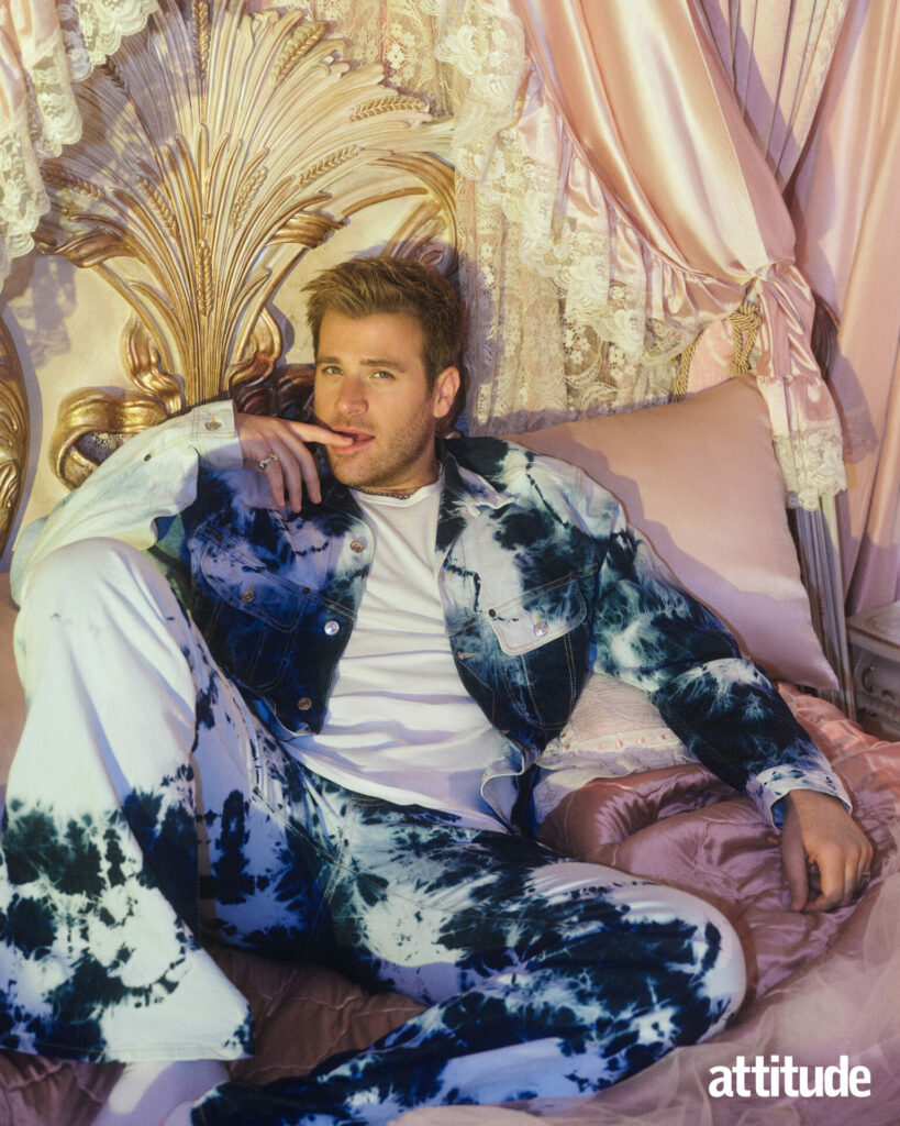 Scott Evans in a tie dye blue and white jacket. He's posing on a regal pink bed with a finder resting on his lips.