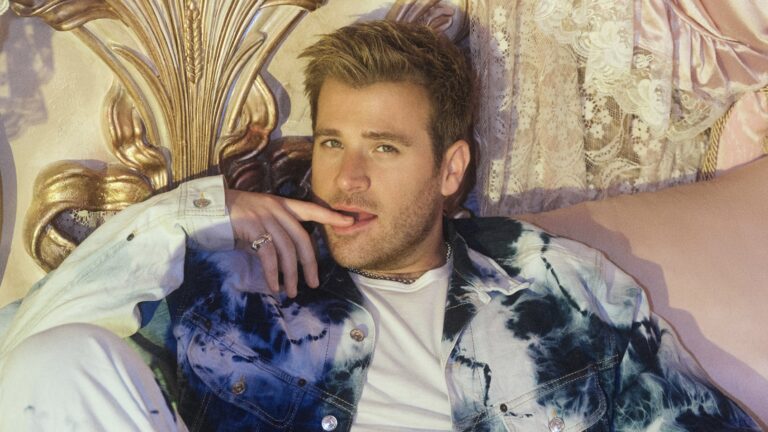 Scott Evans in a tie dye blue and white jacket. He's posing on a regal pink bed with a finder resting on his lips.