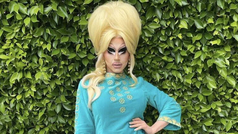 Trixie Mattel standing infant of green shrubbery with a high blonde, full glam and a light blue dress.