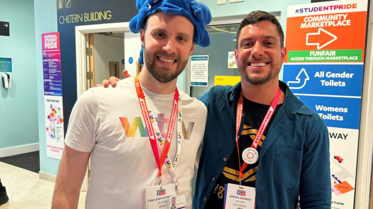 Two men smile into the camera, with one wearing a blue hat