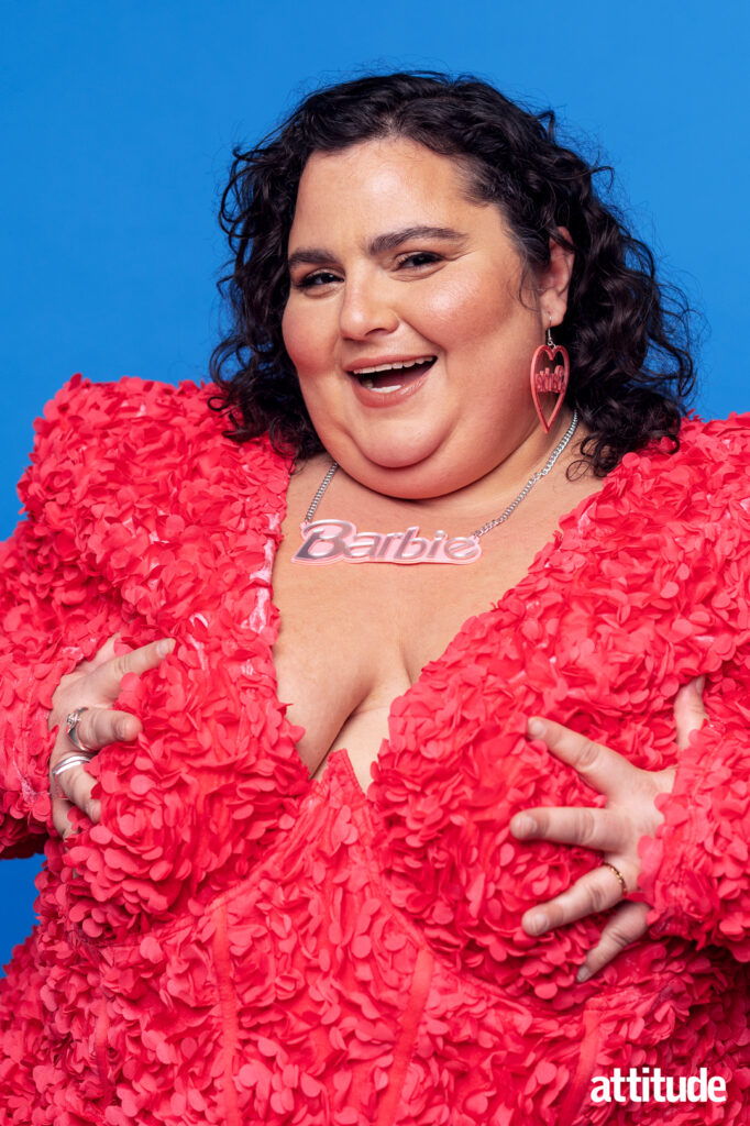 Barbara Butch in a rose-textured dark pink dress with a Barbie necklace. Her hands are pushing against her breasts and she's smiling wide.