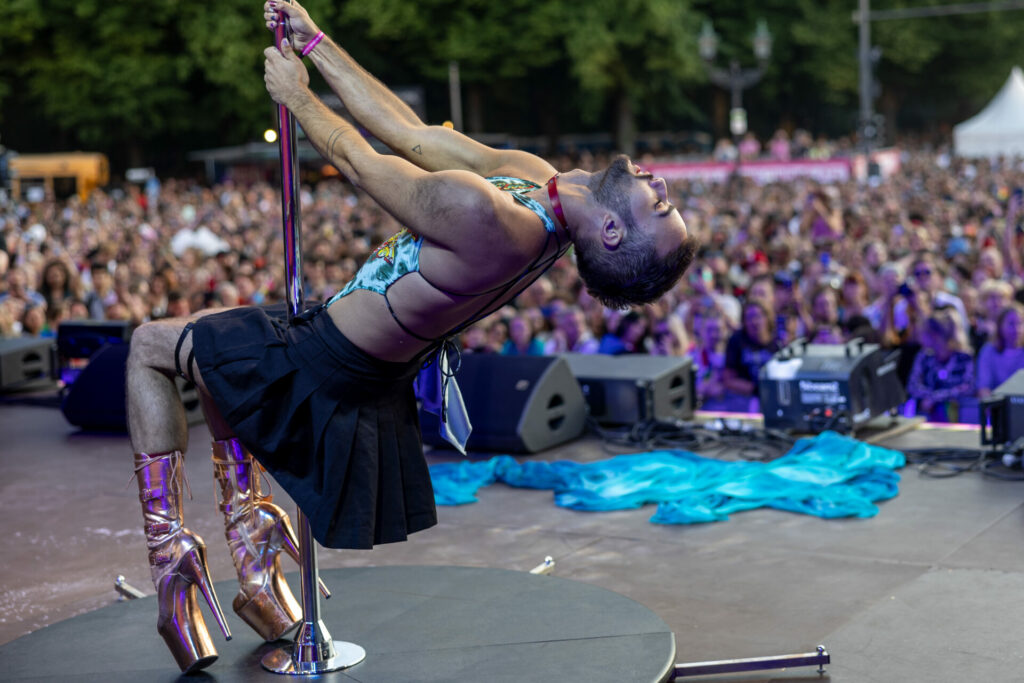 A man in heels spins on a pole on a stage in front of a crowd