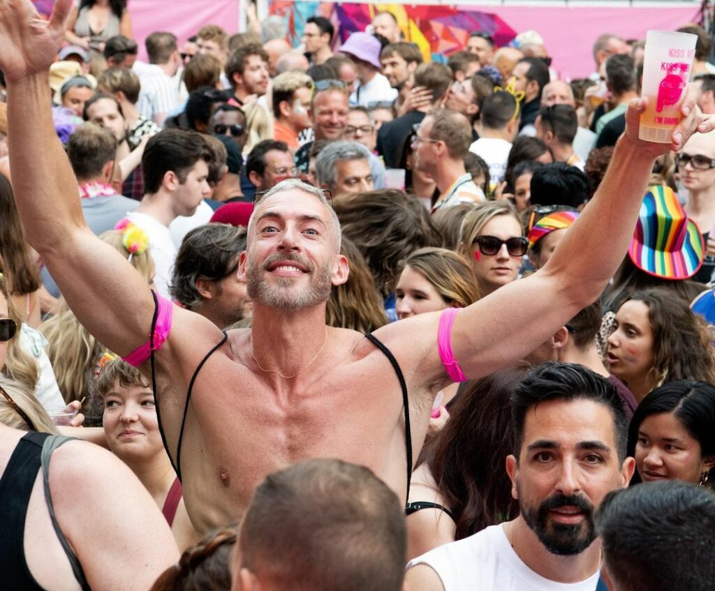 A shirtless man stands with his hands in the air amid a crowd of dancing people