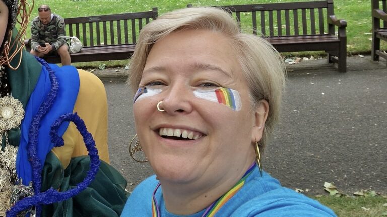 Nancy Kelley with rainbow facepaint. Taking a selfie in a park.