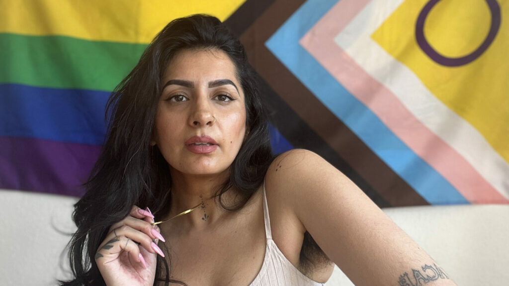 Sharan Dhaliwal posing in front of a Pride Progress flag