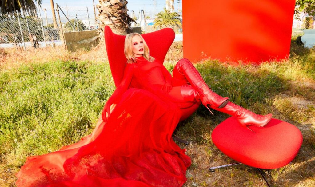 Kylie Minogue sits on a red chair wearing a red gown on a lawn of grass