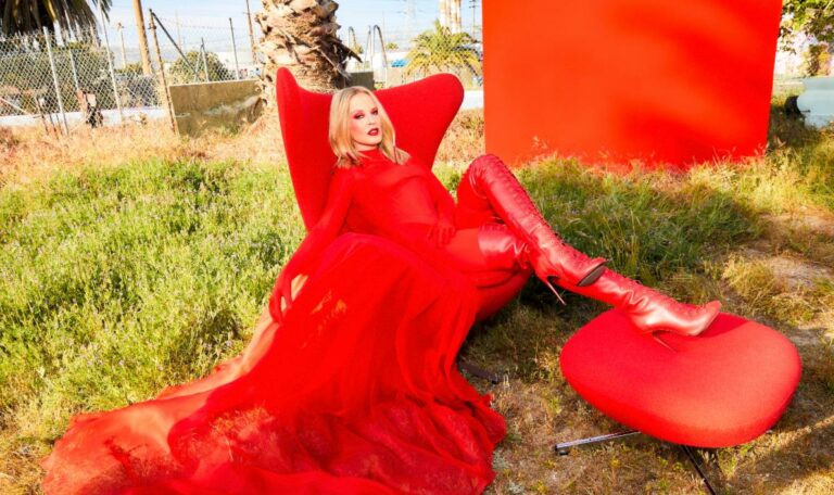 Kylie Minogue sits on a red chair wearing a red gown on a lawn of grass