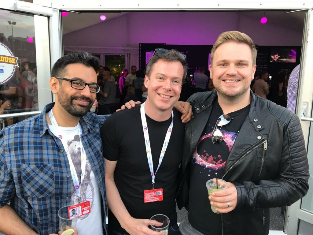 Three men stand together with drinks in their hands looking into the camera