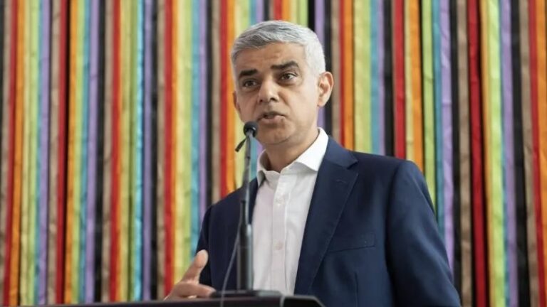 Sadiq Khan in a navy suit against a rainbow-striped background.