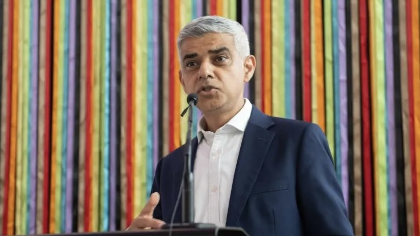 Sadiq Khan in a navy suit against a rainbow-striped background.