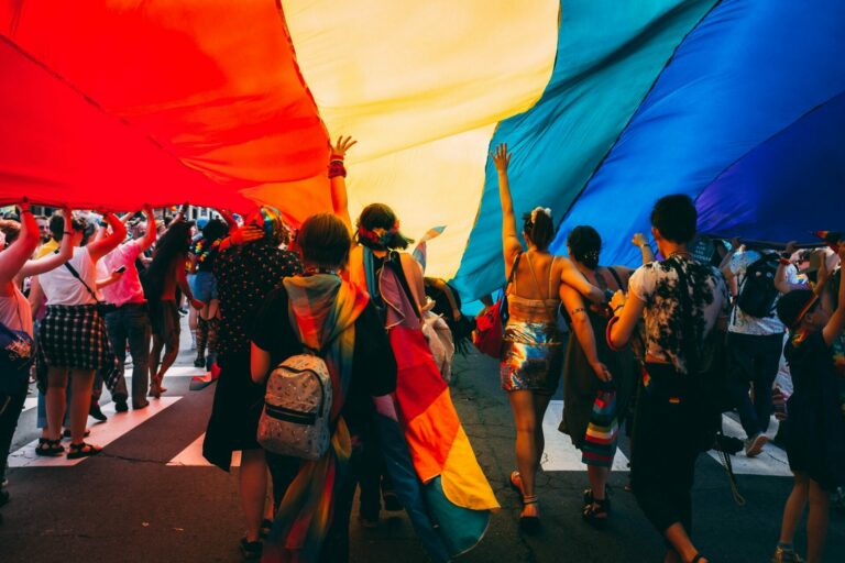 A group of people walk together holding up a huge rainbow flag