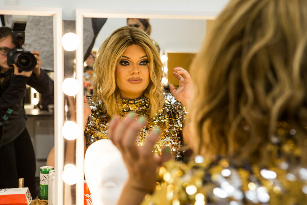 A drag queen sits at a dressing table looking at herself in the mirror