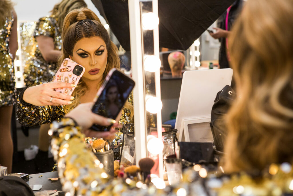A drag queen sits by a makeup mirror and takes a photo of herself with her iPhone