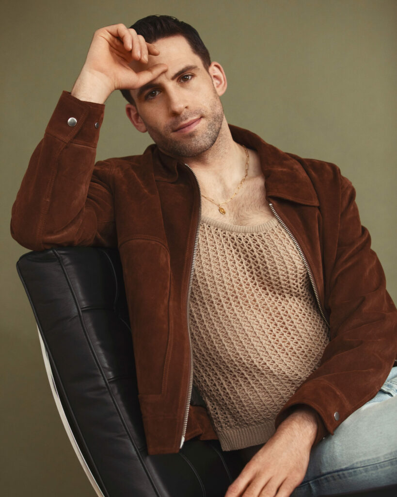 A man sitting on a chair with his hand resting on his face wearing a brown jacket and mesh vest