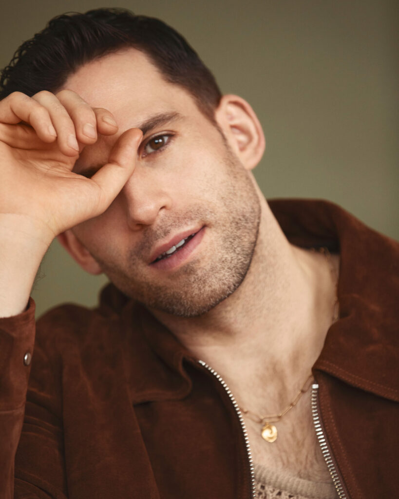 Headshot of a man wearing a brown jacket and mesh vest. He's holding his hand to his face and looking into the camera