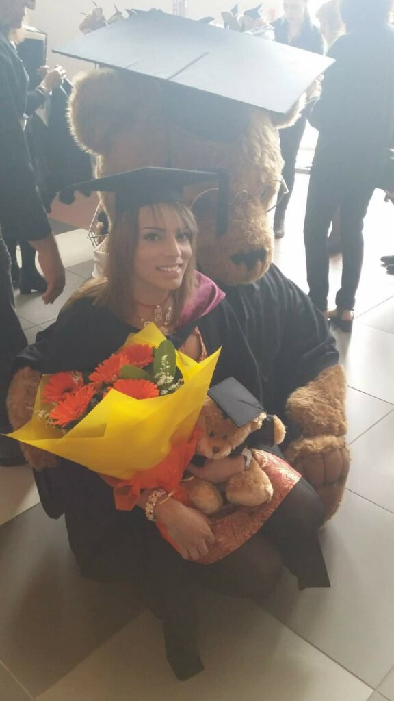 A woman wearing a mortar board hat and black gown holds a bunch of flowers while sitting on a large teddy bear