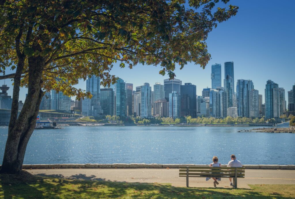 Two people sit on a bench in front of a scene of water and skyscrapers