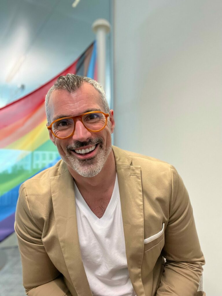 A man wearing a beige blazer and white T-shirt smiles into the camera