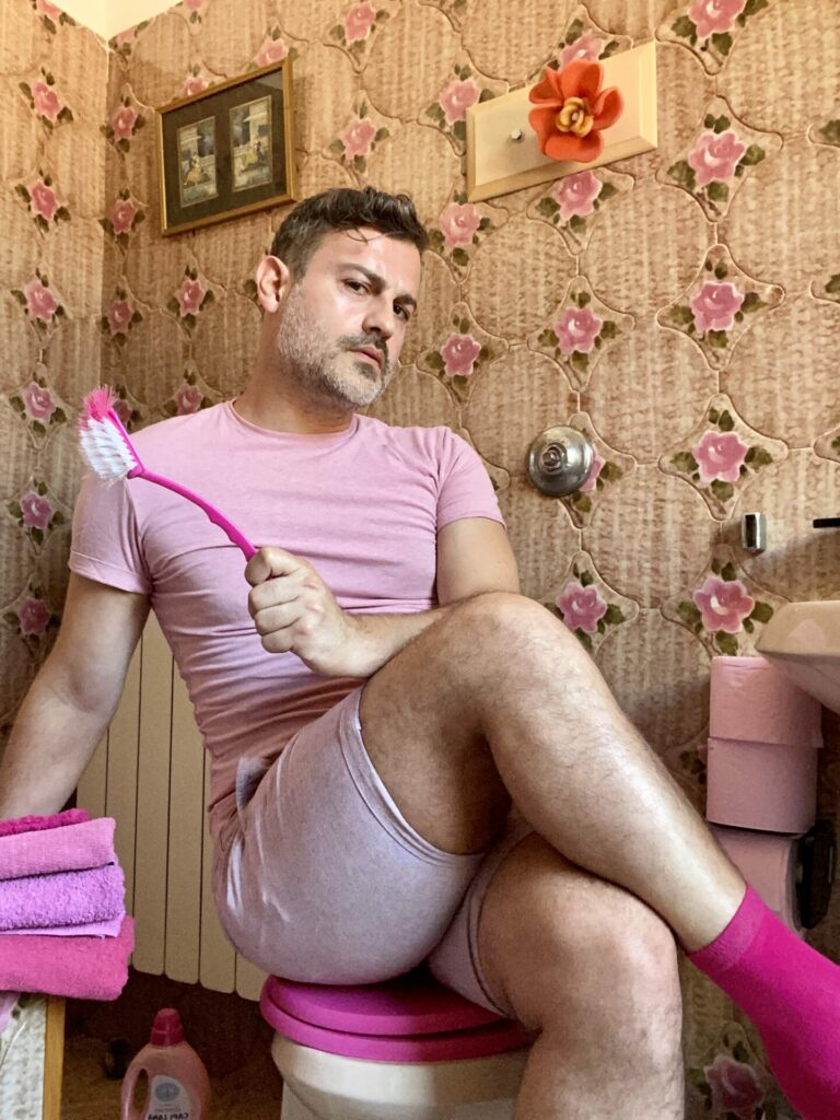 A man wearing a pink T-shirt and pink shorts sits on a toilet holding a pink toilet brush
