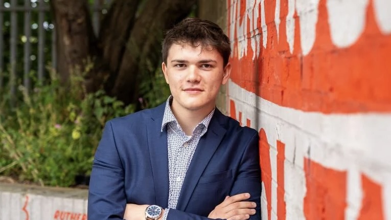 Cameron Eadie, Scottish Green candidate, in a blue suit leaning against a red and white wall