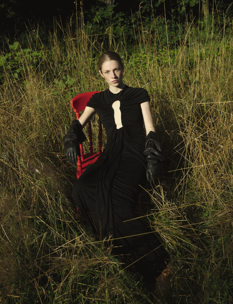 Hunter Schafer sat on a red chair in a black dress and gloves in a field