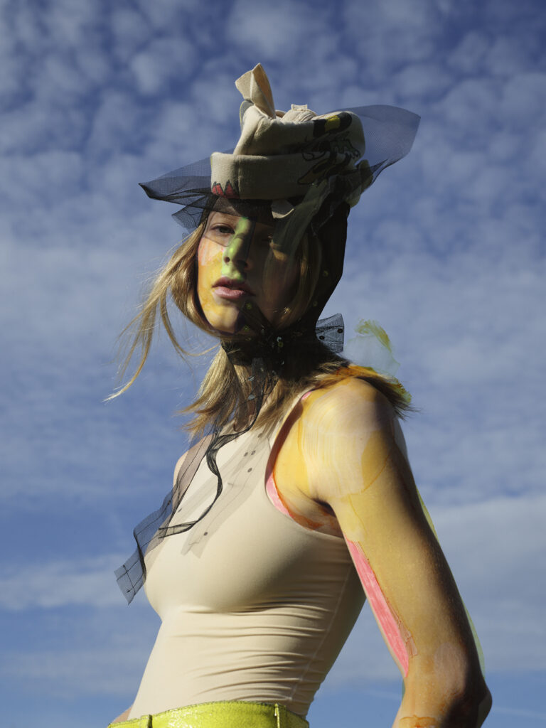Hunter Schafer modeling a hat posing against blue sky. Her arms and face have been coloured