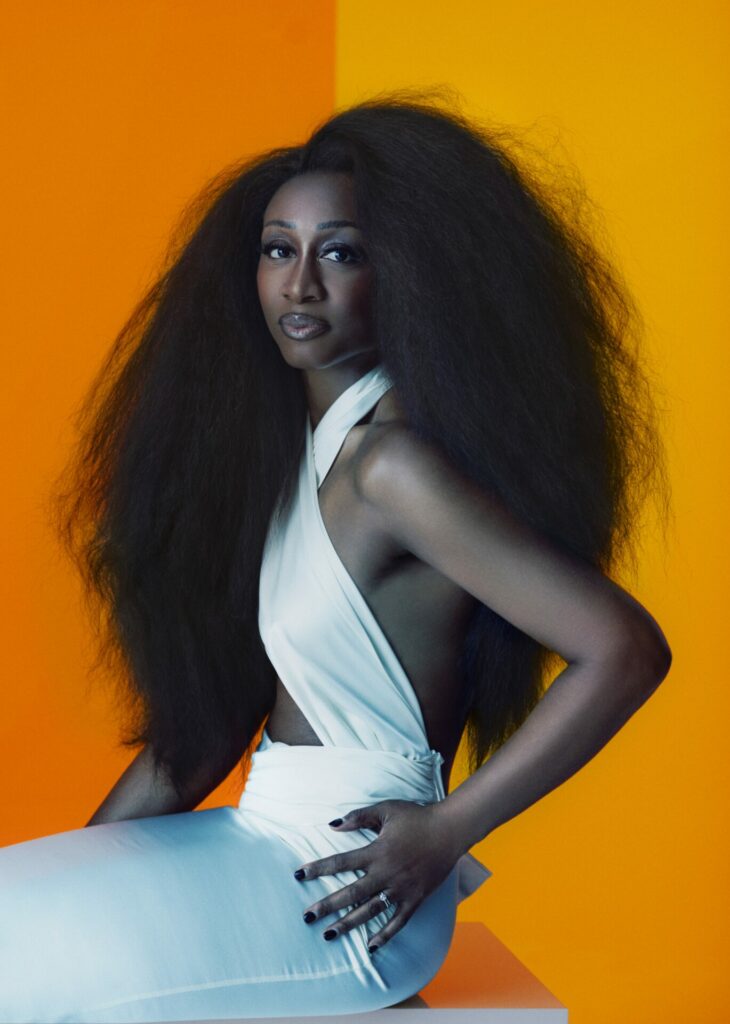 Side view of Beverley Knight wearing a white wrap top and blue skirt against an orange background