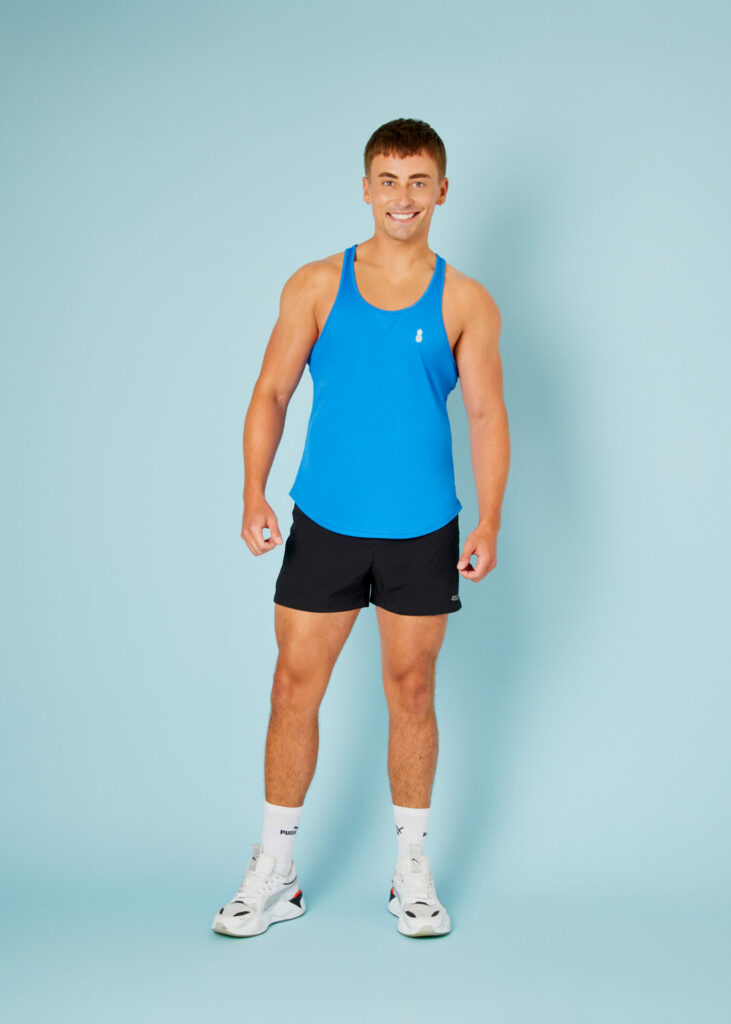 A man wearing a blue vest and black shorts and white socks and trainers stands against a blue background