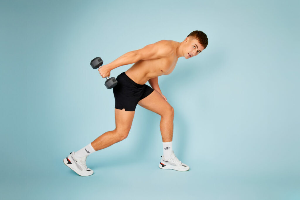 A man wearing black shorts and white trainers and socks holds a dumbell is kneeling