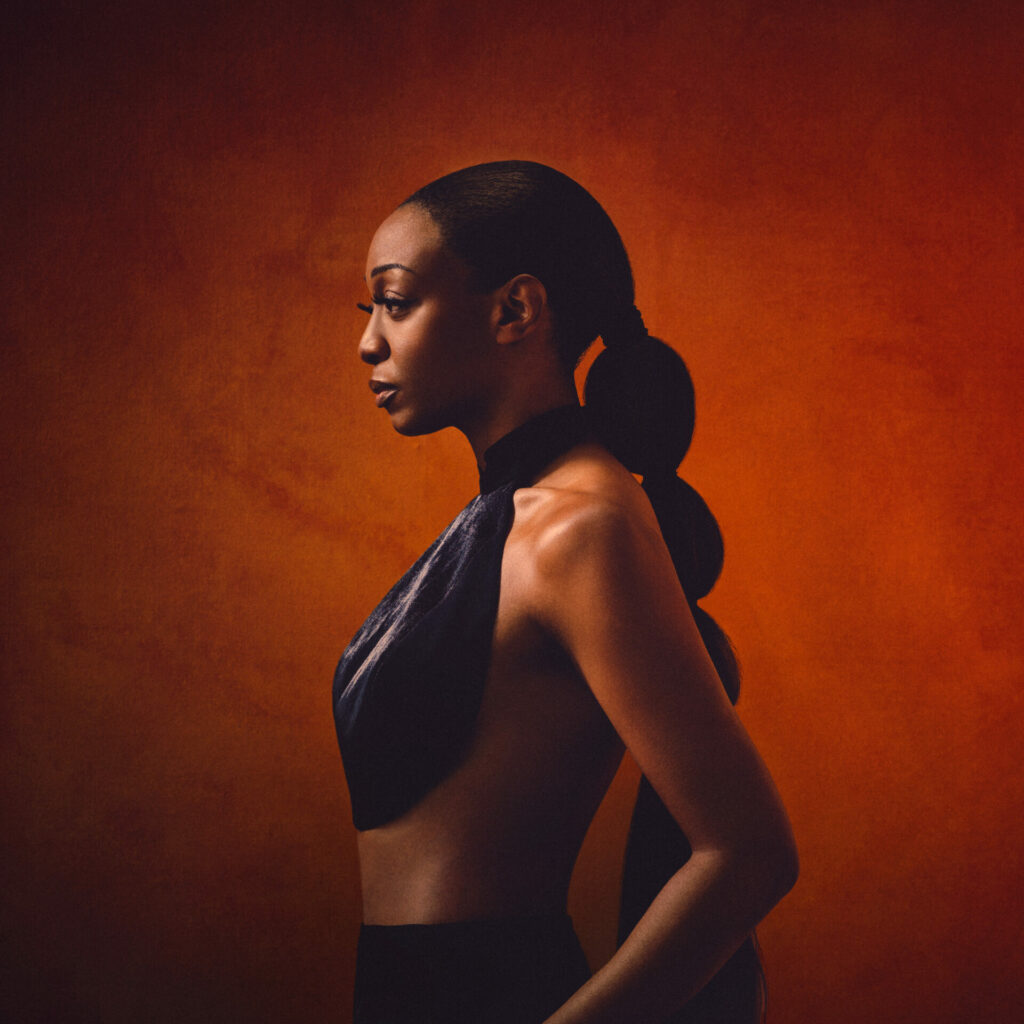 Side view of Beverley Knight wearing a black cropped top and a long plonytail against an orange background
