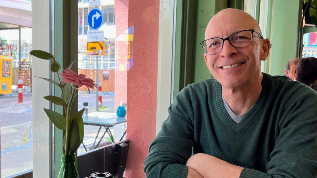 A man sits in a cafe and looks into the camera smiling