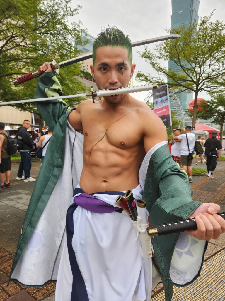 A man with green hair dressed as a samurai with a sword in each hand and a sword in his mouth stands in front of Taipei 101