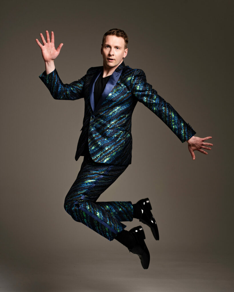 Joe Lycett jumps in the air in a sparkly suit