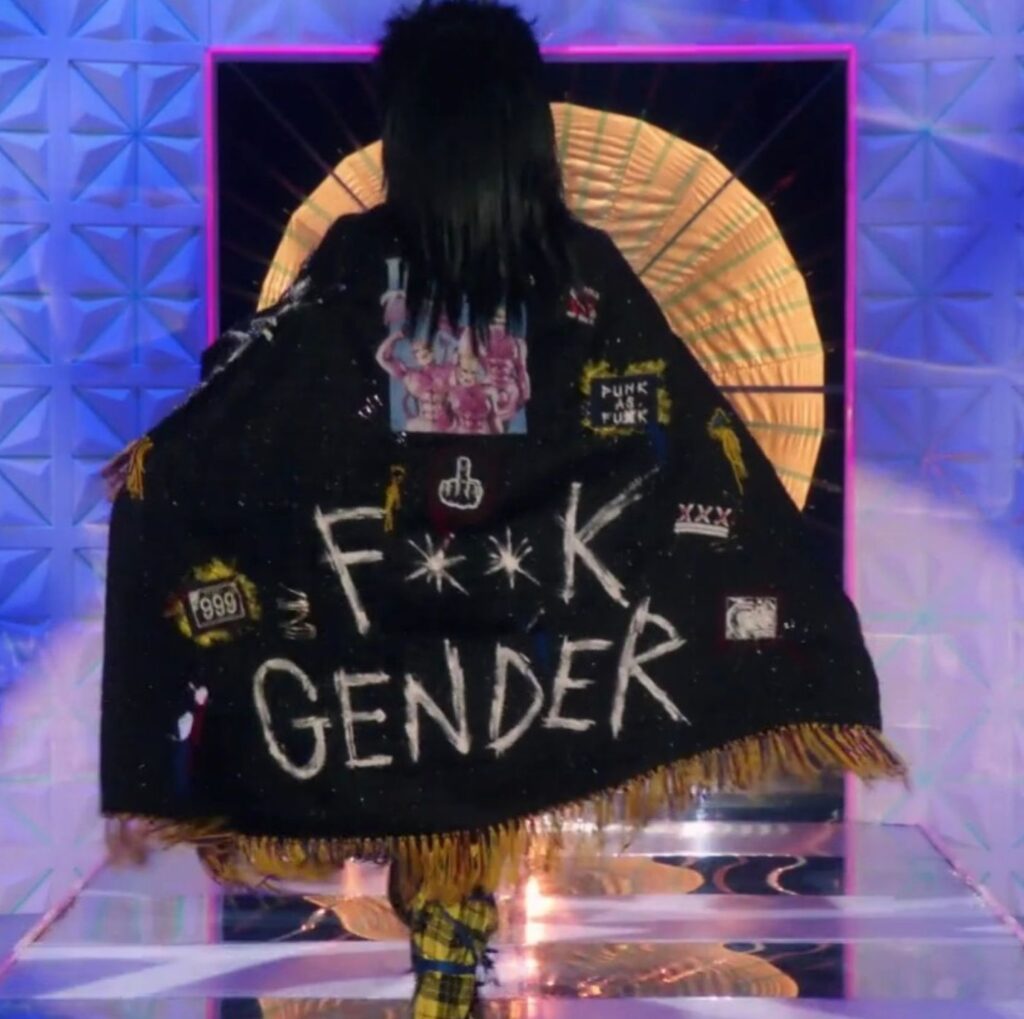 View of Danny Heard from behind wearing a cape on the Drag Race UK runway with the words "F**k gender" on it