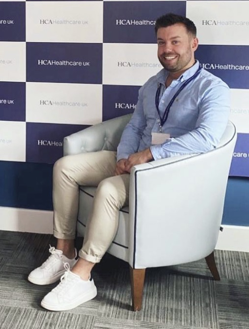 A man sits in front of a chair in front of a board with a company logo